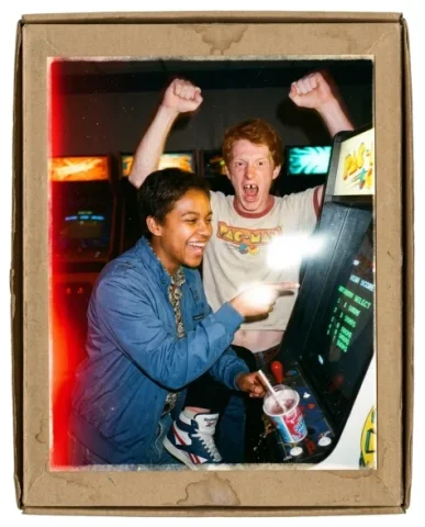 Nostalgic Flash — 80s Arcade Night 3 — Nano Banana AI Prompt: Two friends — a young person with short dark hair and warm brown skin, and a lan...