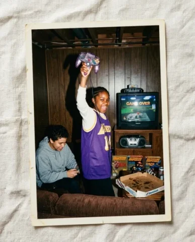 Gaming with [Character] — N64 Era, 1997 3 — Nano Banana AI Prompt: Two friends taking a pizza break on the brown corduroy couch in a wood-paneled b...