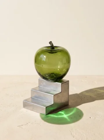 Glass Apple on Aluminum Steps — Nano Banana 2 AI Prompt: An original surreal-but-physically-constructed studio still life: one transparen...