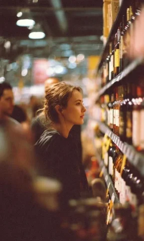 Side-Profile Moment in a Wine Aisle — Nano Banana 2 AI Prompt: A cinematic side-profile portrait of a young woman standing in a grocery store a...