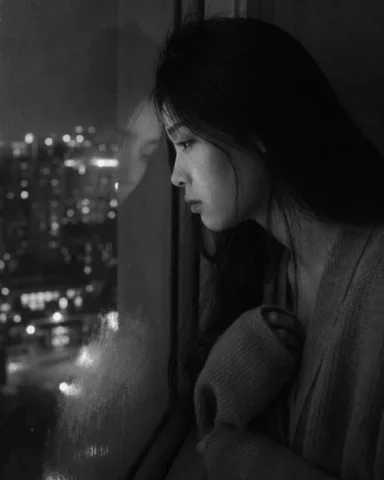 Melancholic Midnight Window Portrait — Nano Banana 2 AI Prompt: Medium close-up portrait, 85mm, a young East Asian woman in her early 20s stands...
