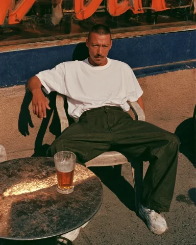 Casual Bar Moment — Nano Banana 2 AI Prompt: A guy in a white tee sitting backwards on a plastic chair outside a bar, beer on...
