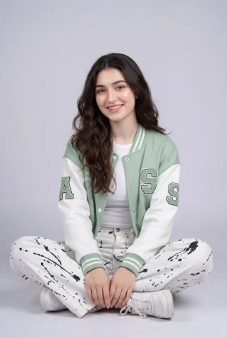 Gray Background Varsity Jacket Cross-Legged Smile Studio Portrait — Nano Banana 2 AI Prompt: Studio portrait of a smiling young woman with long wavy dark brown hair, sitting...