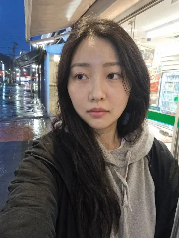 Rainy Convenience Store Selfie — Nano Banana 2 AI Prompt: Create a realistic smartphone front-camera selfie of a young East Asian woman wi...