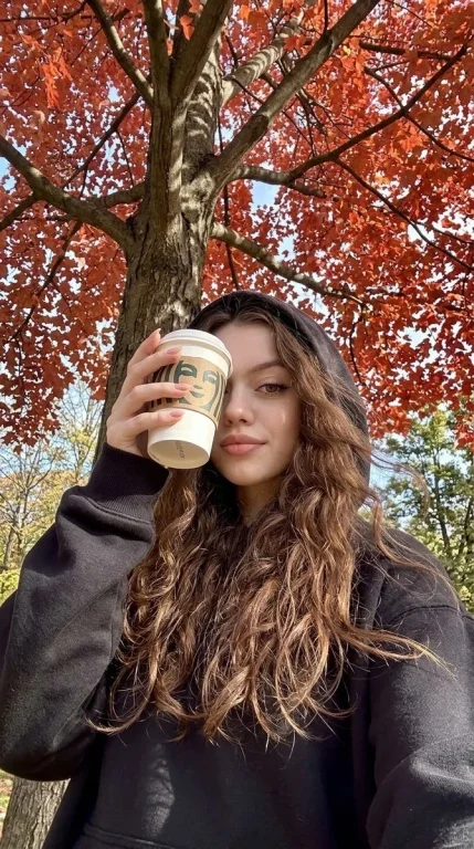 Girl in black hoodie holds coffee under autumn tree