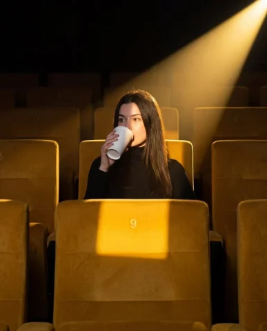 A solitary woman in a dark theater sips from a glowing cup — Nano Banana 2 AI Prompt: A medium shot captures a person sitting alone in an empty movie theater or audit...