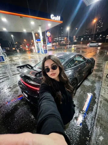 Rainy Night Selfie with Porsche at Urban Gas Station — Nano Banana 2 AI Prompt: Ultra realistic photo of a young woman in the uploaded image at a gas station at...