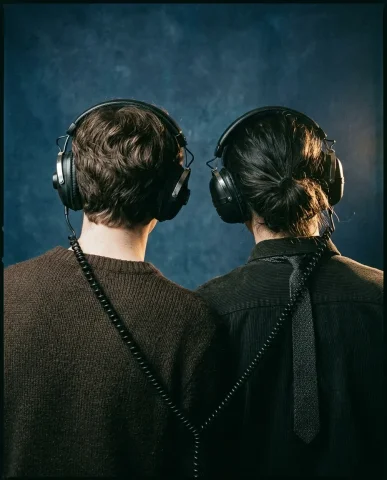 Dual Headphone Portrait 5 — Nano Banana AI Prompt: The deep indigo-blue background stretches above their heads. Warm tungsten light...