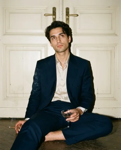 Flash Portrait Against Cream Doors 5 — Nano Banana AI Prompt: The deep midnight-navy double-breasted suit slightly rumpled from the seated pos...